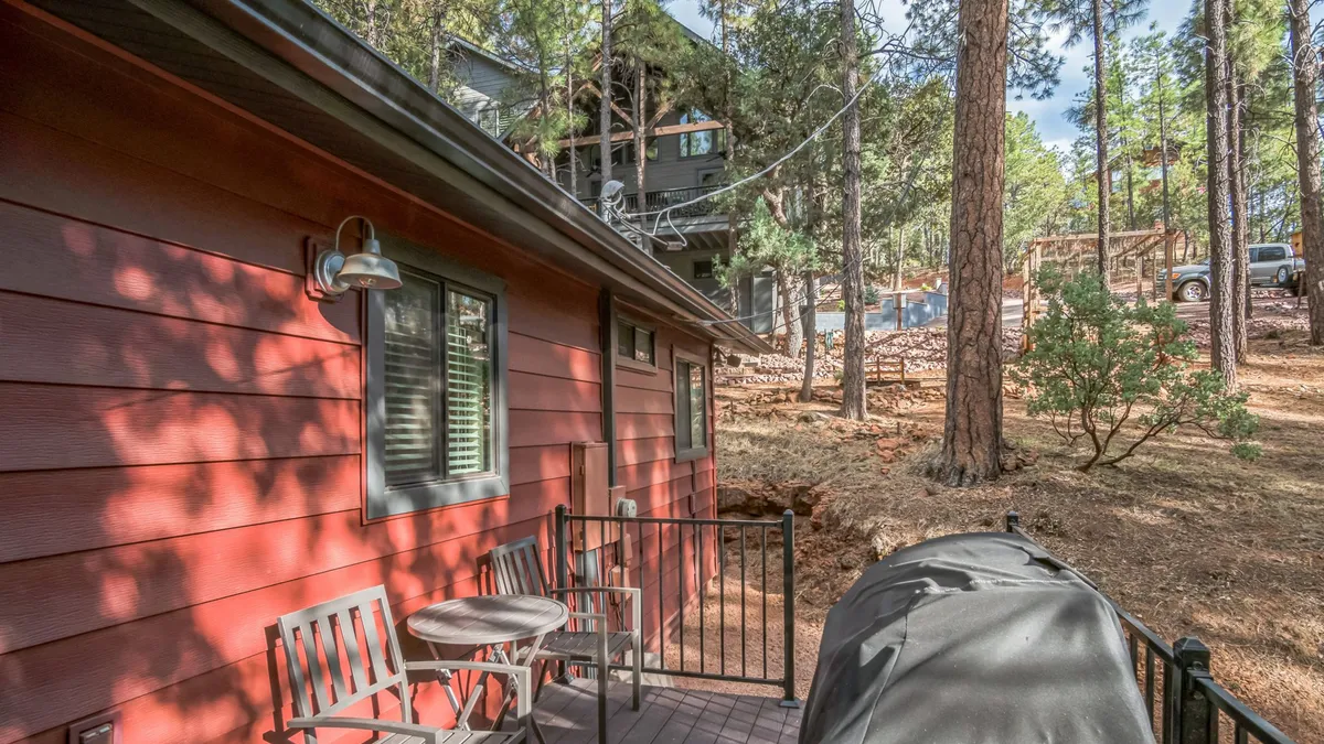 A cozy exterior view featuring red wooden siding, an inviting outdoor seating area, a grill, and tall trees enveloping the patio. The setup is ideal for gatherings or peaceful moments in nature, with plenty of room for pets to relax alongside their owners.