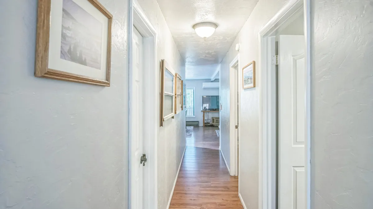 A warmly lit hallway adorned with framed pictures on the walls, with open doorways leading to other rooms and a soft light fixture above. The perspective suggests a sense of movement through the home, hinting at the transition between indoors and the outdoors, and the stories unfolding across seasons.