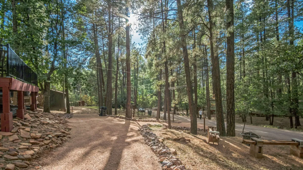 This exterior image features a serene setting with tall pine trees, a gravel path winding through the forest, natural rock formations, and rustic wooden benches for relaxation. The lush forest surrounds the area, highlighting the tranquil, immersive nature experience the property offers.