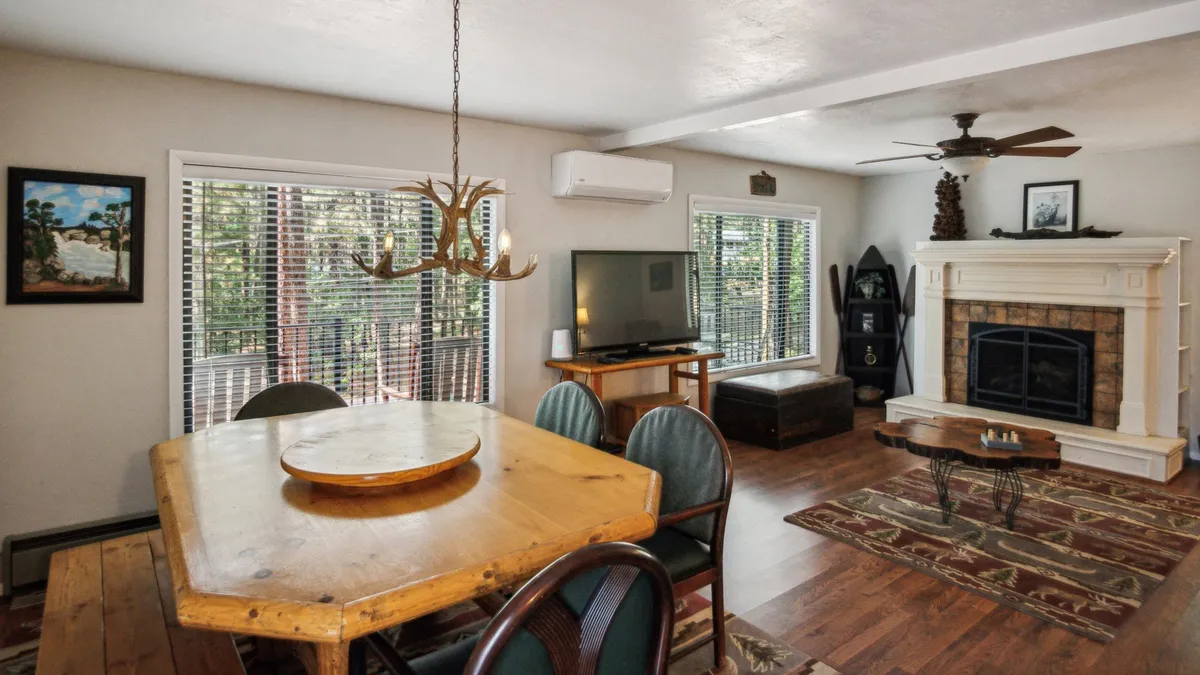 The image highlights a warm, welcoming interior where a dining table sits close to a fireplace, with large windows framing views of nature. A television is visible, reinforcing the space’s multi-functional appeal for meals, movie nights, and relaxation.