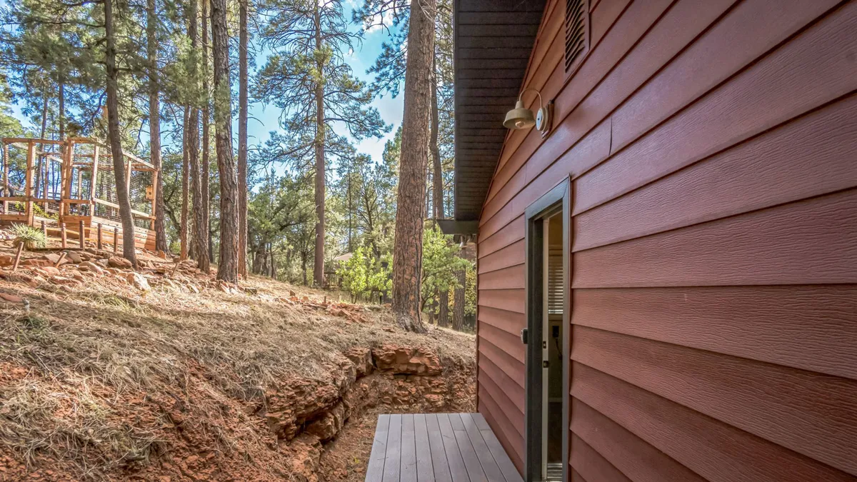 A spacious wooden deck with red exterior walls, bordered by tall pine trees and natural terrain. The deck provides plenty of space for relaxing, while the adjacent yard is ideal for playtime and exploration, making it welcoming for both people and pets.