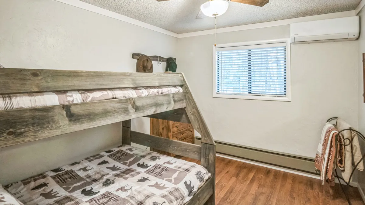 A bright, inviting bedroom featuring a sturdy wooden bunk bed, a window that lets in natural light, a wooden dresser, and a ceiling fan. The atmosphere is relaxed, ideal for sleepovers or quiet mornings, with a view to the outdoors that hints at possible wildlife sightings.