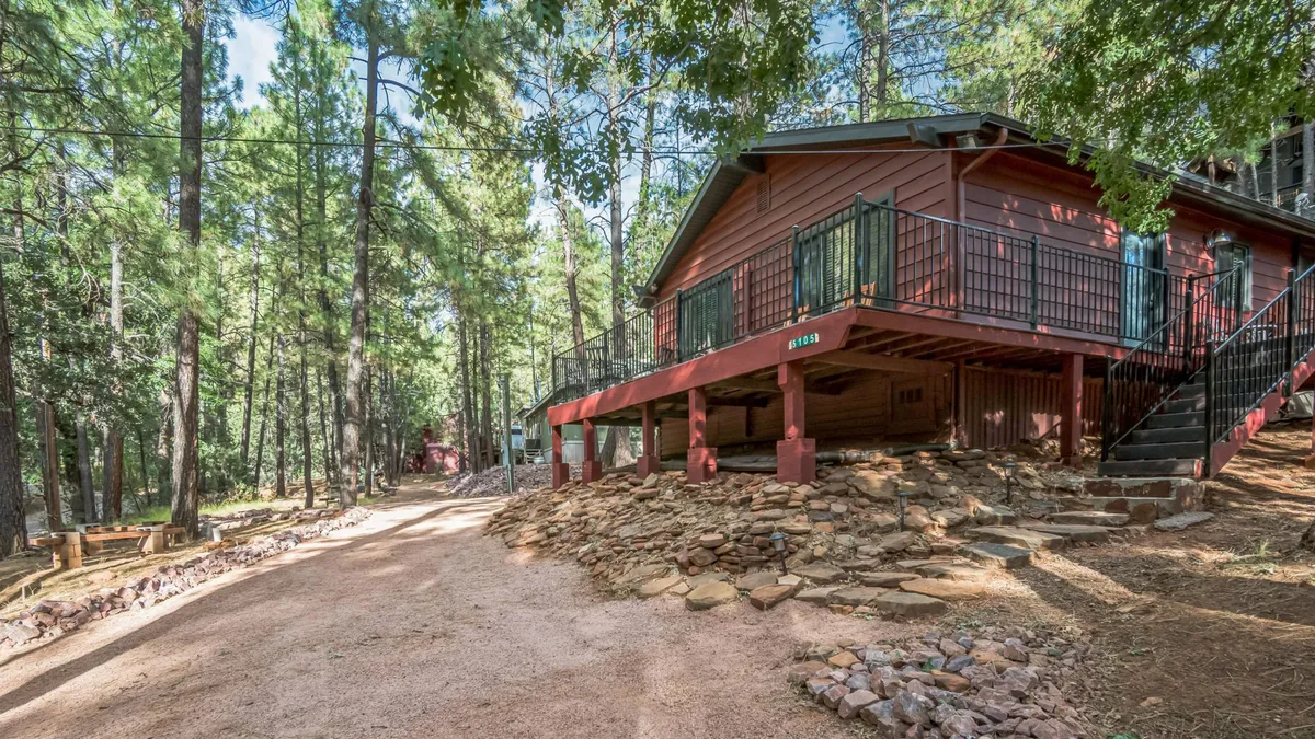 A cozy red wooden cabin sits nestled among towering pine trees, with a gravel pathway leading through natural rocky landscaping. The serene setting hints at peaceful mornings on the porch and evenings under the stars, inviting guests to unwind surrounded by nature.
