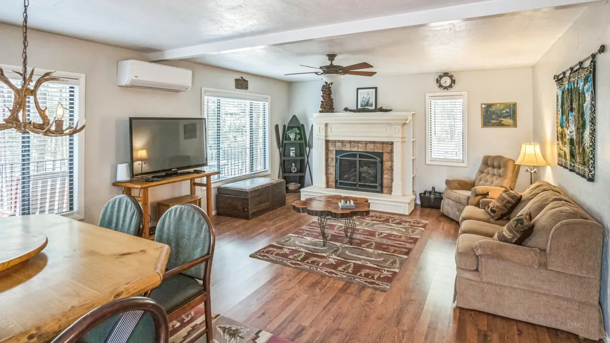 This image presents a cozy living room anchored by a glowing gas fireplace, with a comfortable sofa arranged for conversation or relaxation. A dining table sits nearby, seamlessly blending the living and dining areas. Large windows let in natural light, offering glimpses of the surrounding woodland.