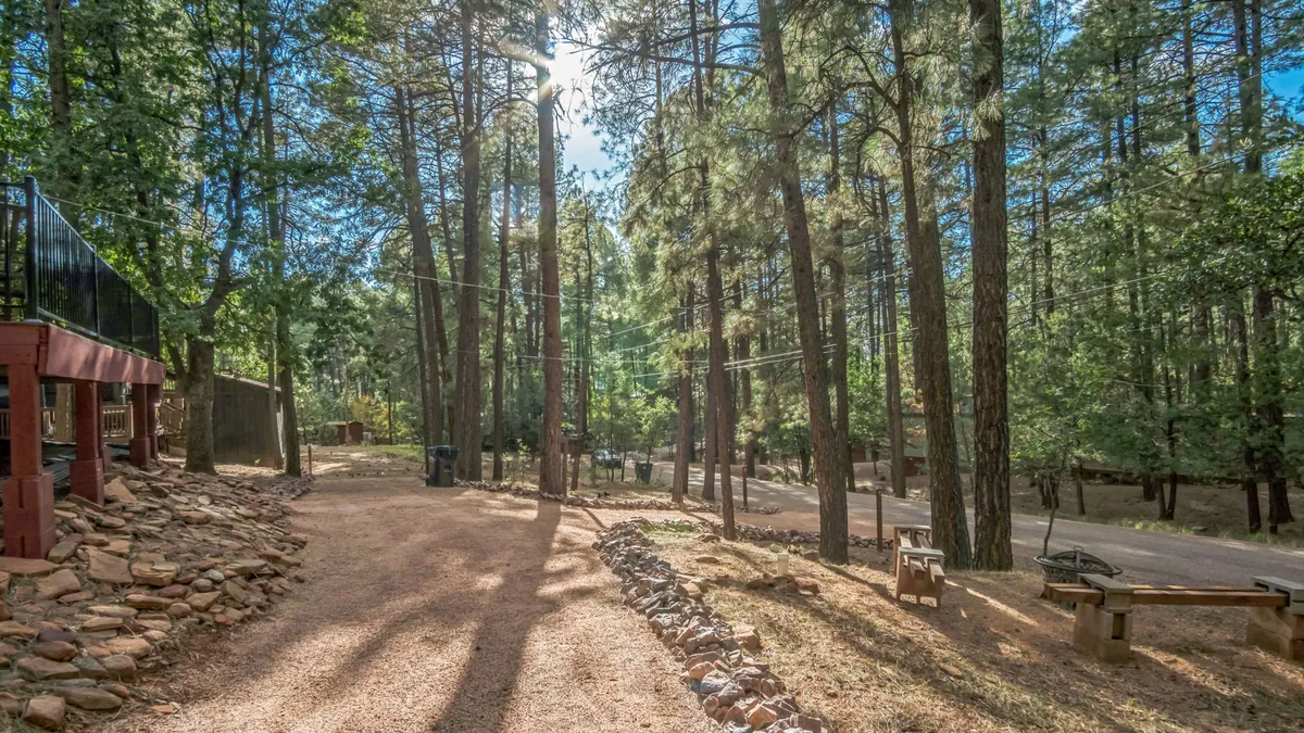 A peaceful outdoor scene featuring tall pine trees, a gravel path lined with rock borders, and a cozy seating area. The landscape suggests opportunities for hiking, stargazing, and quiet relaxation among the trees.